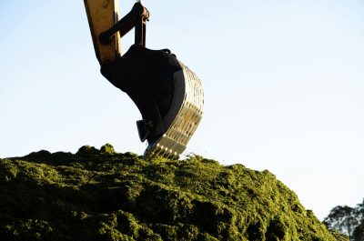 pexels-photo-27434717-27434717 An excavator digging and moving a large pile of green organic material in outdoor daylight.